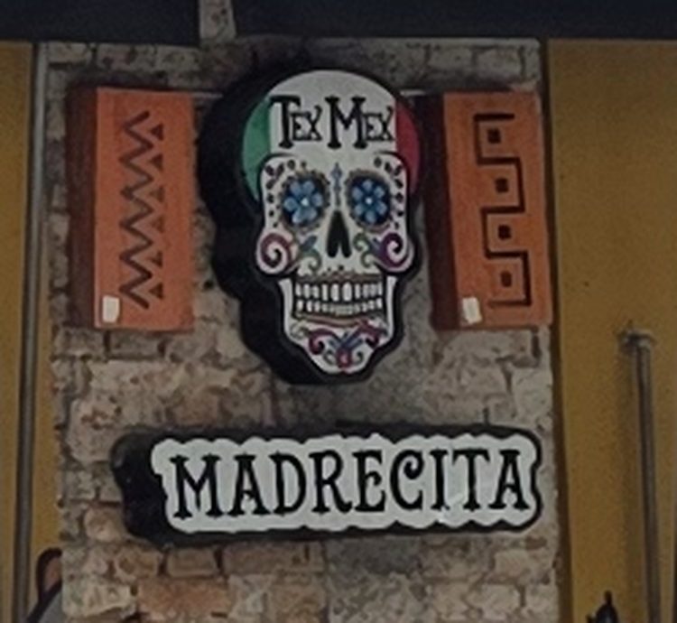 Placa em forma de caveira mexicana colorida com a inscrição Tex-Mex, sobre o letreiro Madrecita, entre dois ladrilhos de terracota.