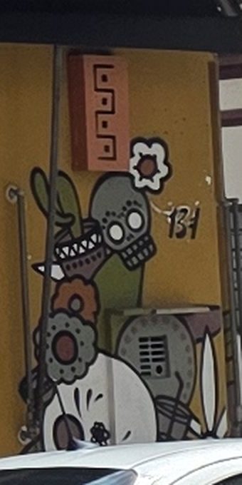 Mural na lateral da porta: calavera verde, cacto e flores laranja, abaixo de um ladrilho de terracota com grecas.