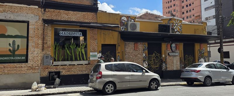 Fachada da Madrecita: casa antiga de tijolo pintada de amarelo, com toldo preto, letreiro Madrecita na vitrine cheia de cactos, portas de enrolar desenhadas e murais de calaveras e flores.