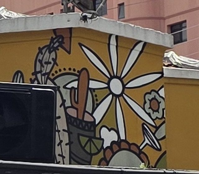 Mural no alto da fachada amarela: margarida branca gigante, cacto num copo e fatia de limão.