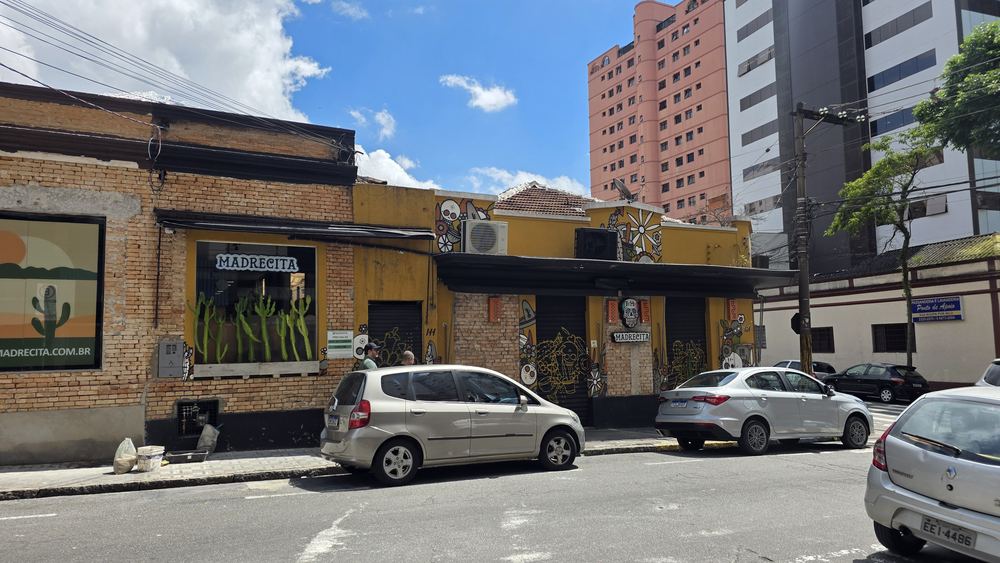 Vista da rua: a casa amarela da Madrecita com o toldo preto, ao lado de um prédio rosa e de um prédio de vidro.